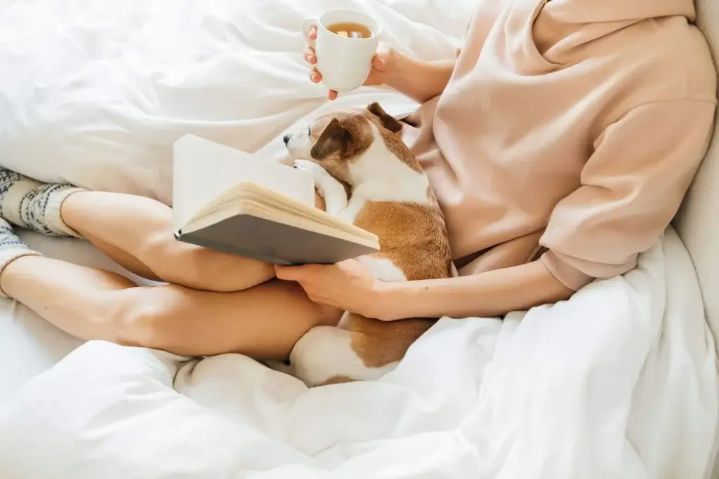 dog in lap of woman drinking coffee and reading book in bed
